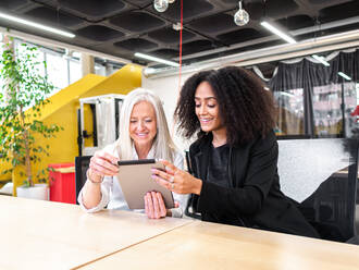 Positive multirassische weibliche Mitarbeiter in formeller Kleidung sitzen am Tisch und surfen auf einem Tablet während der Zusammenarbeit an einem Projekt - ADSF43101