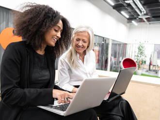 Positive junge afroamerikanische Frau mit Afrofrisur sitzt auf einer Bank im Büro und tippt auf einem Netbook, während eine ältere Führungskraft einen Computer benutzt - ADSF43108