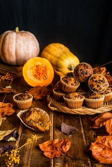 Komposition aus appetitlichen Muffins mit Schokolade-Chips auf Holztisch unter verstreuten Herbstblätter und Kürbisse auf dunklem Hintergrund platziert - ADSF43170