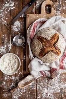 Draufsicht auf einen unordentlichen rustikalen Holztisch aus Weizenmehl und ein Brett mit frisch gebackenem Sauerteigbrotlaib - ADSF43173