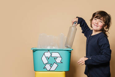 Glückliches Mädchen in schwarzem Hemd, das lächelnd in die Kamera schaut und eine Flasche in eine Box mit Recycling-Symbol an einer beigen Wand stellt - ADSF43190