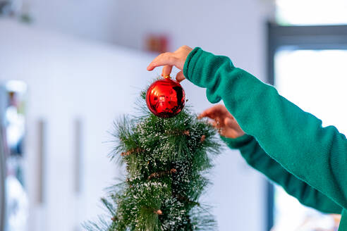 Seitenansicht eines abgeschnittenen, nicht erkennbaren Kindes mit Händen im Pullover, das eine rote Christbaumkugel an den Weihnachtsbaum hängt, während es sich auf die Weihnachtsfeier zu Hause vorbereitet - ADSF43198