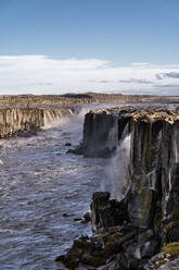 Luftaufnahme des Selfoss-Wasserfalls, der von einer felsigen Klippe in den Fluss Jokulsa Fjollum fällt, unter blauem Himmel mit Sonnenlicht in Island - ADSF43208