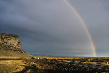 Atemberaubende Landschaft von Regenbogen in bewölktem Himmel über trockenen grasbewachsenen Wiese und Asphaltstraße, die zu Lomagnupur felsigen Berg in vulkanischem Gelände in Island - ADSF43225