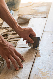 From above of crop anonymous male master holding metal piece and leveling tile during renovation on sunny day - ADSF43230