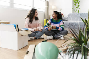 Happy young friends sitting with coffee cup on floor at home - JCZF01174