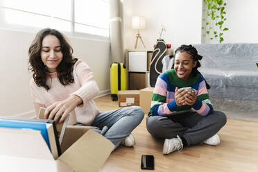 Smiling young woman unpacking box with friend holding coffee cup at home - JCZF01177