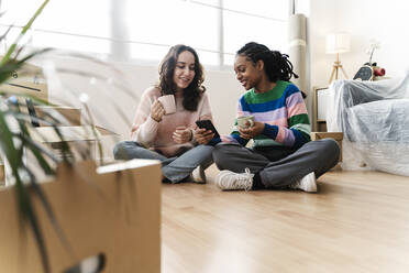 Smiling young woman sharing smart phone sitting on floor at home - JCZF01179