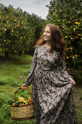 Glückliche rothaarige Frau steht mit Korb vor Orangenbäumen - NJAF00213