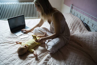 Arbeitende Mutter, die einen Laptop benutzt und mit einem kleinen Jungen spielt, der zu Hause auf dem Bett liegt - NJAF00227