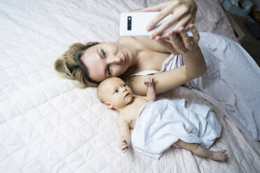 Mother taking selfie with baby boy through smart phone on bed at home - NJAF00232