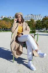 Glückliche junge Frau mit Baskenmütze sitzt auf einem Stuhl im Park - KIJF04519