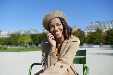 Happy young woman wearing beret sitting on chair in front of blue sky - KIJF04521