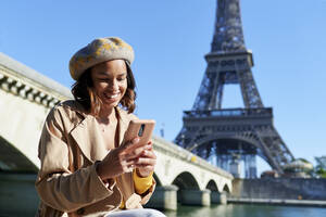 Glückliche junge Frau, die mit ihrem Smartphone vor dem Eiffelturm eine SMS schreibt, Paris, Frankreich - KIJF04539