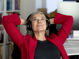 Lächelnde Geschäftsfrau mit drahtlosen Kopfhörern, die mit den Händen hinter dem Kopf sitzt - FLLF00843