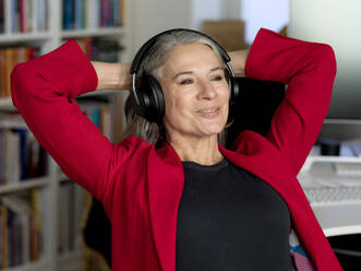 Happy businesswoman wearing wireless headphones sitting with hands behind head - FLLF00844