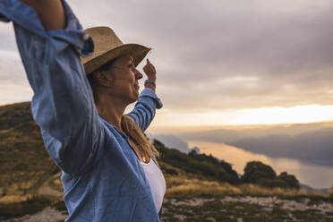 Carefree mature woman with arms raised standing under sky at sunset - UUF28208