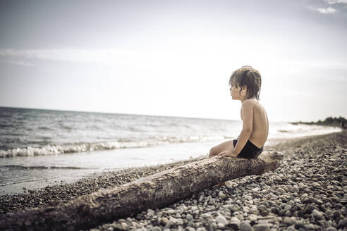 Hemdloser Junge sitzt auf einem Baumstamm am Strand - ANAF00986