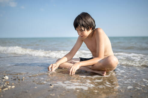 Junge spielt im nassen Sand und sitzt im Schneidersitz am Strand - ANAF00987