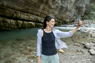 Smiling woman taking selfie through smart phone standing near riverbank - ANAF00995