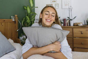 Glückliche Frau mit geschlossenen Augen umarmt Kissen im Bett zu Hause - TYF00716