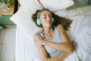 Lächelnde Frau mit geschlossenen Augen, die über Kopfhörer im Bett Musik hört - TYF00721