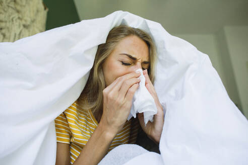 Woman wiping nose with tissue under blanket at home - TYF00729