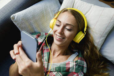 Happy woman using mobile phone wearing headphones lying on couch at home - TYF00738