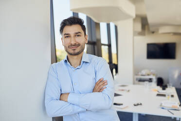 Confident businessman with arms crossed leaning on wall at workplace - BSZF02062