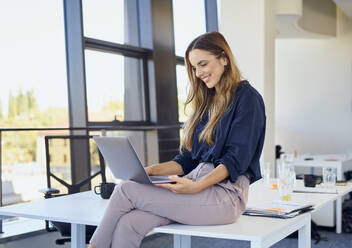 Smiling businesswoman using laptop sitting on desk at workplace - BSZF02076