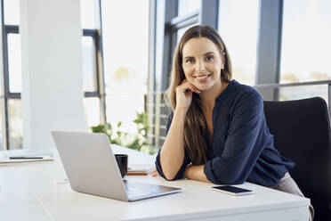 Smiling businesswoman leaning on desk by laptop at office - BSZF02083