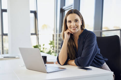 Lächelnde Geschäftsfrau, die sich im Büro mit dem Laptop auf den Schreibtisch lehnt - BSZF02083