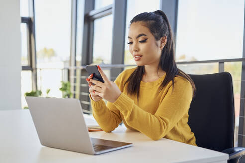 Junge Geschäftsfrau, die im Büro ein Mobiltelefon am Laptop benutzt - BSZF02089