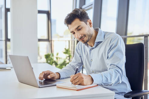 Businessman taking down notes in diary by laptop at office - BSZF02090