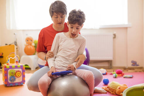 Physiotherapeutin und Mädchen mit Behinderung sitzen auf einem Fitnessball in einem Rehabilitationszentrum - OSF01383