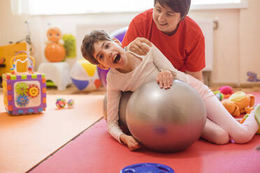 Glückliche Physiotherapeutin und Mädchen mit Behinderung trainieren auf einem Fitnessball - OSF01384