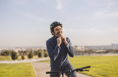 Happy mature man adjusting helmet on sunny day - JCCMF09335