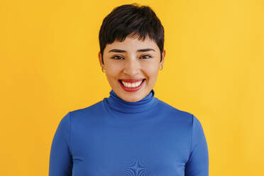 Happy young woman with pixie haircut standing against yellow background - TCEF02228