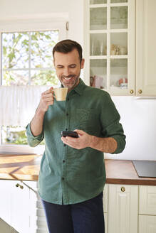Lächelnder Mann telefoniert beim Kaffeetrinken in seiner Küche - BSZF02164