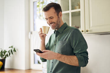 Lächelnder Mann telefoniert beim Kaffeetrinken in seiner Küche - BSZF02165