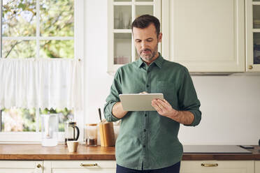 Reifer Mann mit digitalem Tablet in der Küche stehend - BSZF02178