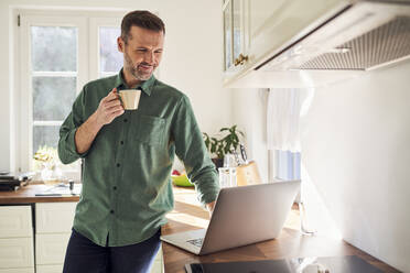 Lächelnder Mann, der einen Laptop benutzt und in der Küche Kaffee trinkt - BSZF02180