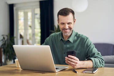 Cheerful man paying with smartphone on laptop sitting at table at home - BSZF02189