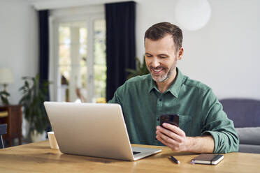 Fröhlicher Mann, der mit seinem Smartphone am Laptop bezahlt, der zu Hause am Tisch sitzt - BSZF02190