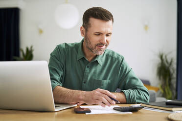 Mann zu Hause sitzend mit Laptop und Dokumenten, die die Hausfinanzen prüfen - BSZF02192