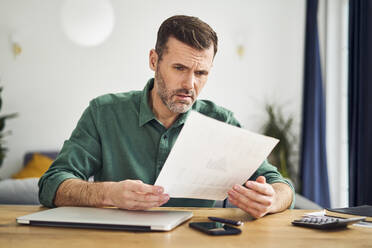 Upset man looking at document with financial figuers while sitting at table at home - BSZF02197