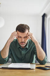 Worried man looking at document with financial figuers while sitting at table at home - BSZF02198