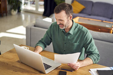 Smiling man working from home using laptop holding documents - BSZF02201