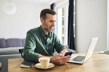 Selbstbewusster Mann sitzt zu Hause am Tisch und benutzt einen Laptop, um mit dem Handy zu bezahlen - BSZF02216