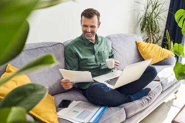 Glücklicher Mann, der von zu Hause aus arbeitet und sich Dokumente ansieht, während er mit seinem Laptop auf dem Sofa sitzt und Kaffee trinkt - BSZF02241
